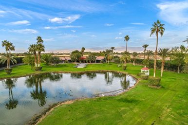 Desert oasis estate with palm trees, manicured lawn, white gazebo and a reflecting pond with ducks, mountains and blue sky