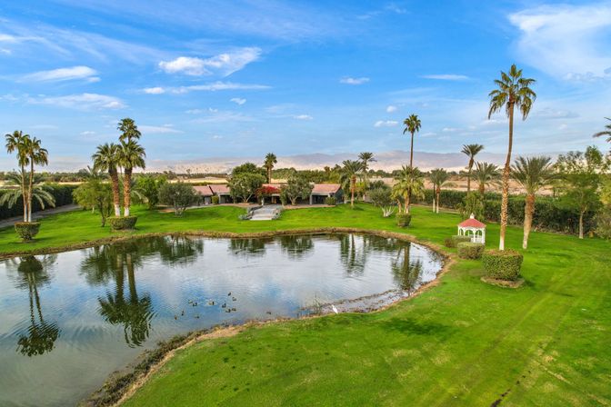 Desert oasis estate with palm trees, manicured lawn, white gazebo and a reflecting pond with ducks, mountains and blue sky