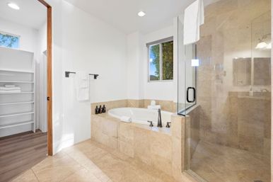 Spa-like modern master bathroom with beige stone tile, built-in soaking tub, glass-enclosed walk-in shower, matte black fixtures, white towels and open linen shelves illuminated by natural window light.
