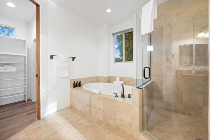Spa-like modern master bathroom with beige stone tile, built-in soaking tub, glass-enclosed walk-in shower, matte black fixtures, white towels and open linen shelves illuminated by natural window light.