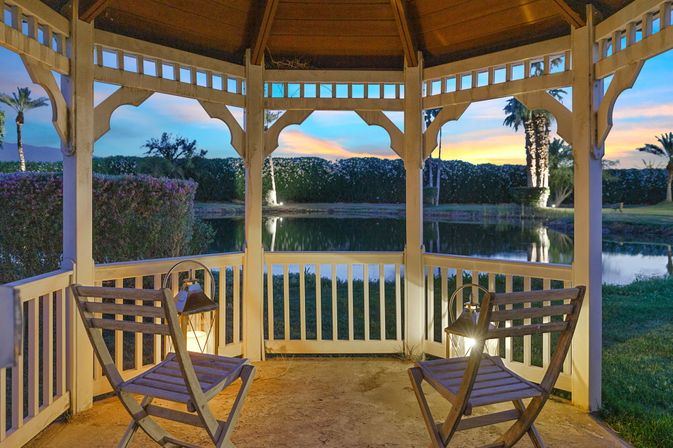 Cozy wooden gazebo at sunset with two folding chairs and lanterns overlooking a reflective pond and palm trees