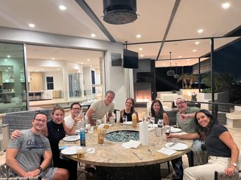 Smiling group of eight friends enjoying a patio dinner around a round stone table with a glass fire-pit, wine glasses and plates on a modern covered terrace with a fireplace, palm trees and city lights visible at night