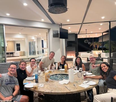 Group of friends gathered around a round fire‑pit dining table on a modern covered patio at night, with wine glasses, plates, a lit fireplace and city lights visible beyond the glass walls.