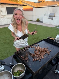 Energetic woman in a white dress laughing while flipping diced steak on a large flat-top griddle with a spatula and fork at a backyard cookout
