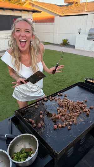 Energetic woman in a white dress laughing while flipping diced steak on a large flat-top griddle with a spatula and fork at a backyard cookout