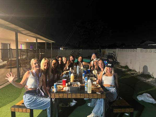 Nighttime backyard pool party: a group of women seated at a long outdoor picnic table on artificial turf, smiling and enjoying plates of food, drinks and conversation under patio lights.