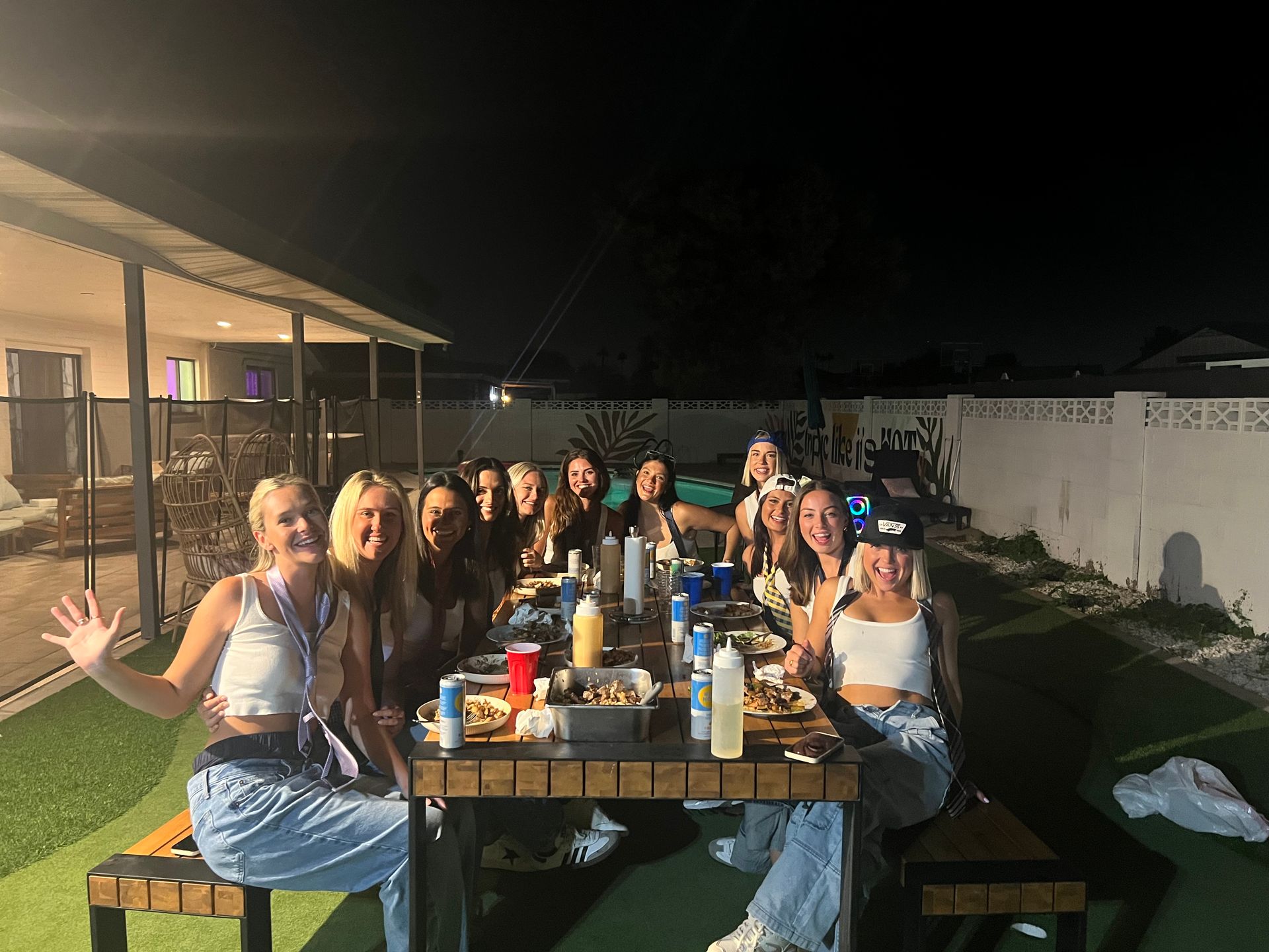 Backyard pool party at night: cheerful group of friends seated around a long outdoor dining table with plates, drinks and condiments, smiling under patio lights beside a swimming pool and fenced yard.