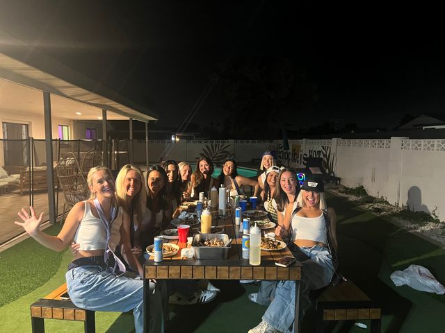 Backyard pool party at night: cheerful group of friends seated around a long outdoor dining table with plates, drinks and condiments, smiling under patio lights beside a swimming pool and fenced yard.