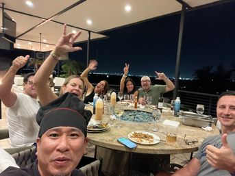 Nighttime rooftop dinner patio: group of friends around a round table with a glass fire pit, wine bottles, plates and city lights on the horizon, selfie-style with a person in a black chef hat in the foreground and others waving and giving thumbs-up.