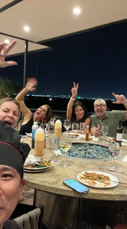 Friends cheering at a rooftop patio dinner at night with city skyline lights in the background, raised hands, wine glasses and shared plates on a round table