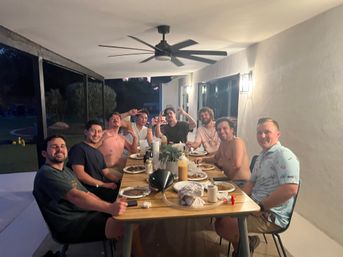 Group of friends smiling around a long wooden table for a casual backyard patio dinner at dusk, plates, condiments and drinks visible under a ceiling fan.