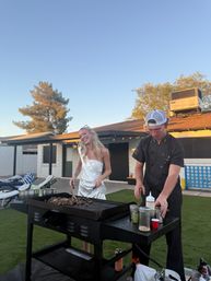 Smiling woman in a white dress and sash beside a chef in a cap grilling chopped steak on a large flat-top griddle at a residential backyard patio during golden hour, outdoor party vibes.