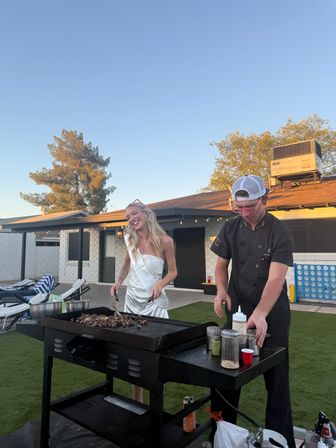 Smiling woman in a white dress and sash beside a chef in a cap grilling chopped steak on a large flat-top griddle at a residential backyard patio during golden hour, outdoor party vibes.