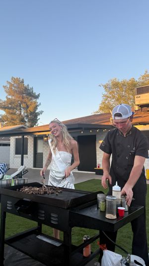 Smiling woman in a white dress and sash beside a chef in a cap grilling chopped steak on a large flat-top griddle at a residential backyard patio during golden hour, outdoor party vibes.