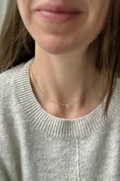 Close-up of a person's neck and lower face wearing a delicate gold chain with a small round clear pendant resting above a light gray knit sweater, minimalist jewelry and cozy style.