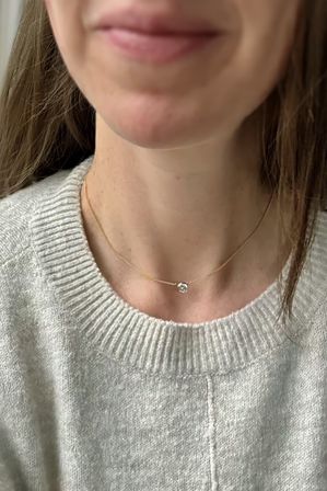 Close-up of a person's neck and lower face wearing a delicate gold chain with a small round clear pendant resting above a light gray knit sweater, minimalist jewelry and cozy style.