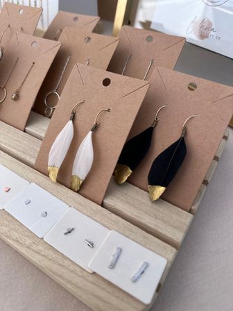 Boho handmade feather earrings with gold-dipped tips — white and black pairs mounted on kraft cards on a wooden display rack with small minimalist stud earrings, artisan market jewelry display.