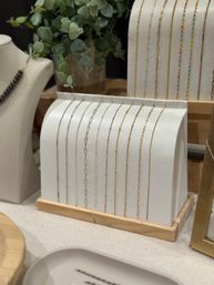 Dainty gold and silver chain necklaces neatly draped over a curved white display with a wooden base, styled with eucalyptus greenery in a boutique jewelry countertop scene.