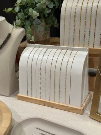 Dainty gold and silver chain necklaces neatly draped over a curved white display with a wooden base, styled with eucalyptus greenery in a boutique jewelry countertop scene.