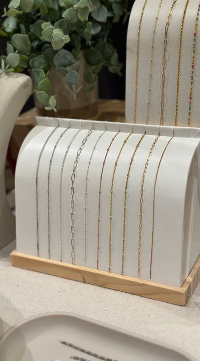Dainty gold and silver chain necklaces neatly draped over a curved white display with a wooden base, styled with eucalyptus greenery in a boutique jewelry countertop scene.