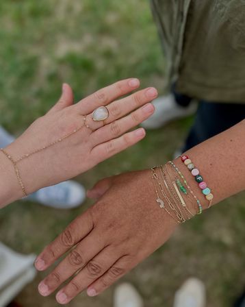 Close-up outdoors over grass showing two hands: one with a gold chain hand bracelet and moonstone ring, the other with stacked gold chains and colorful bead name bracelet, boho jewelry style.