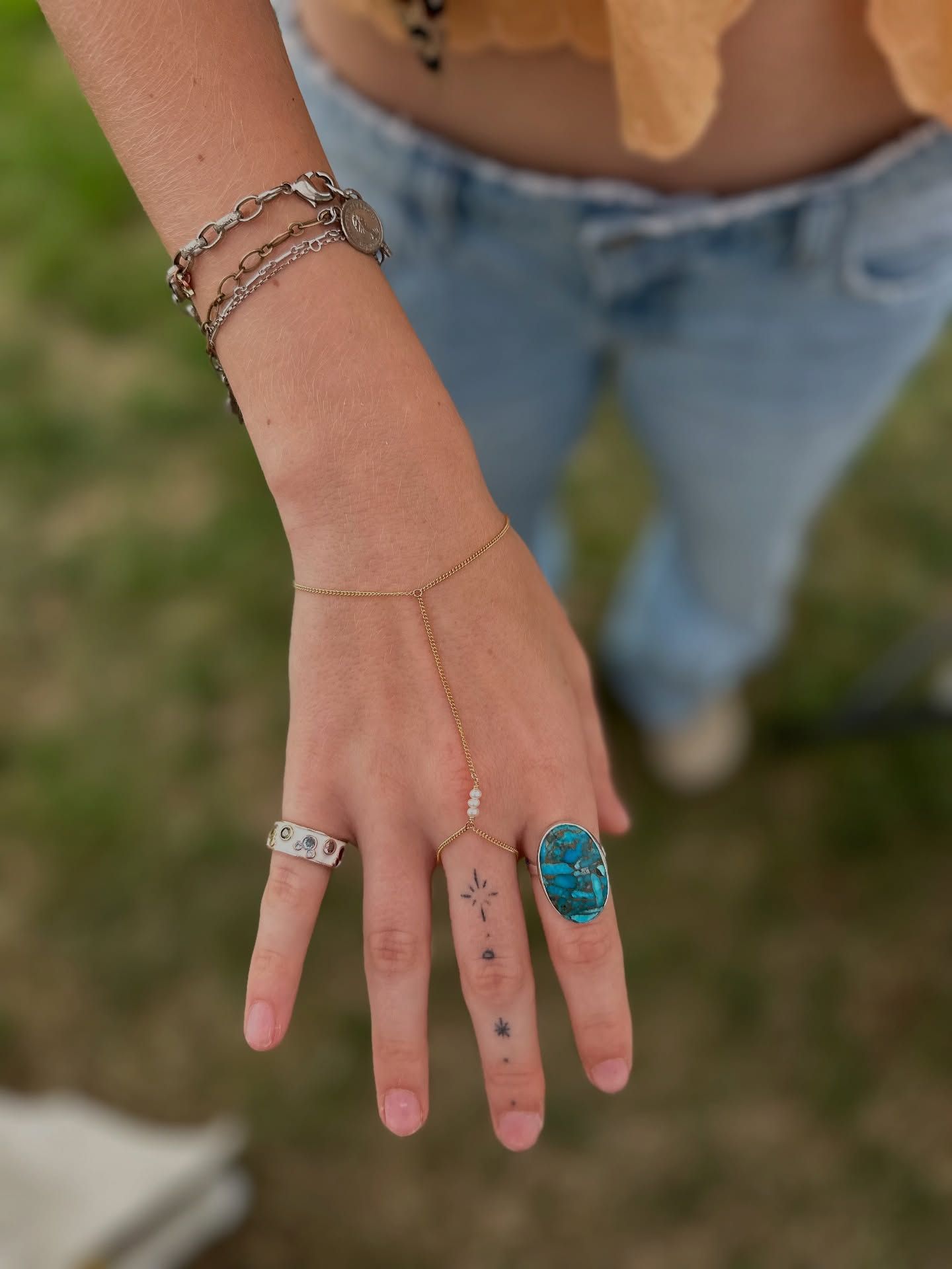 Close-up of a hand with boho jewelry — large turquoise statement ring, silver band, delicate gold hand-chain with tiny pearls, stacked chain bracelets and small star finger tattoos against a blurred grassy outdoor background and casual jeans.