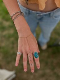 Close-up of a hand with boho jewelry — large turquoise statement ring, silver band, delicate gold hand-chain with tiny pearls, stacked chain bracelets and small star finger tattoos against a blurred grassy outdoor background and casual jeans.