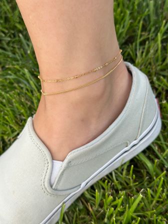 Close-up of an ankle wearing two delicate gold anklets above a pale green slip-on canvas shoe on a green lawn