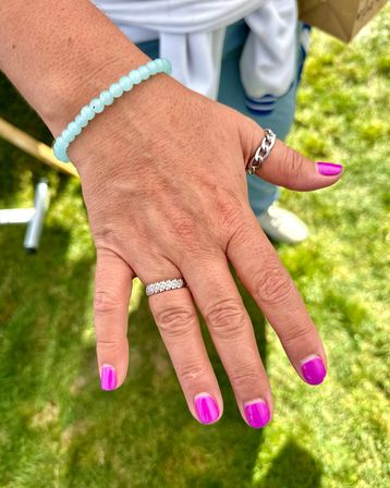 Close-up of a hand over green lawn featuring bright fuchsia nail polish, a diamond-studded band on the ring finger, a chunky silver chain thumb ring and a pale aqua beaded bracelet.
