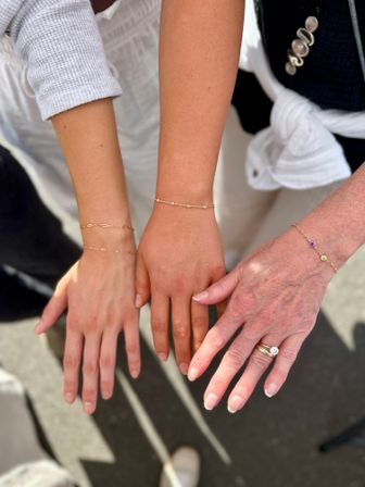 Close-up of three sunlit wrists wearing delicate gold chain bracelets and a diamond ring, hands extended over pavement in an outdoor setting.