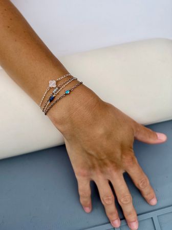 Close-up of a tanned wrist wearing stacked delicate silver chain bracelets with a small blue gemstone and pale pink charm, against a white cushion and gray surface.