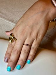 Close-up of a hand with bright turquoise manicure and a glitter accent nail, wearing a chunky gold sculptural ring and a delicate gemstone chain ring on a neutral surface