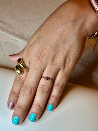 Close-up of a hand with bright turquoise manicure and a glitter accent nail, wearing a chunky gold sculptural ring and a delicate gemstone chain ring on a neutral surface