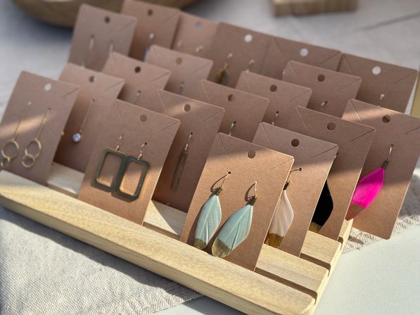 Sunlit display of handmade earrings on kraft cards in a wooden rack — feather, geometric and minimalist dangle styles at an outdoor artisan market.