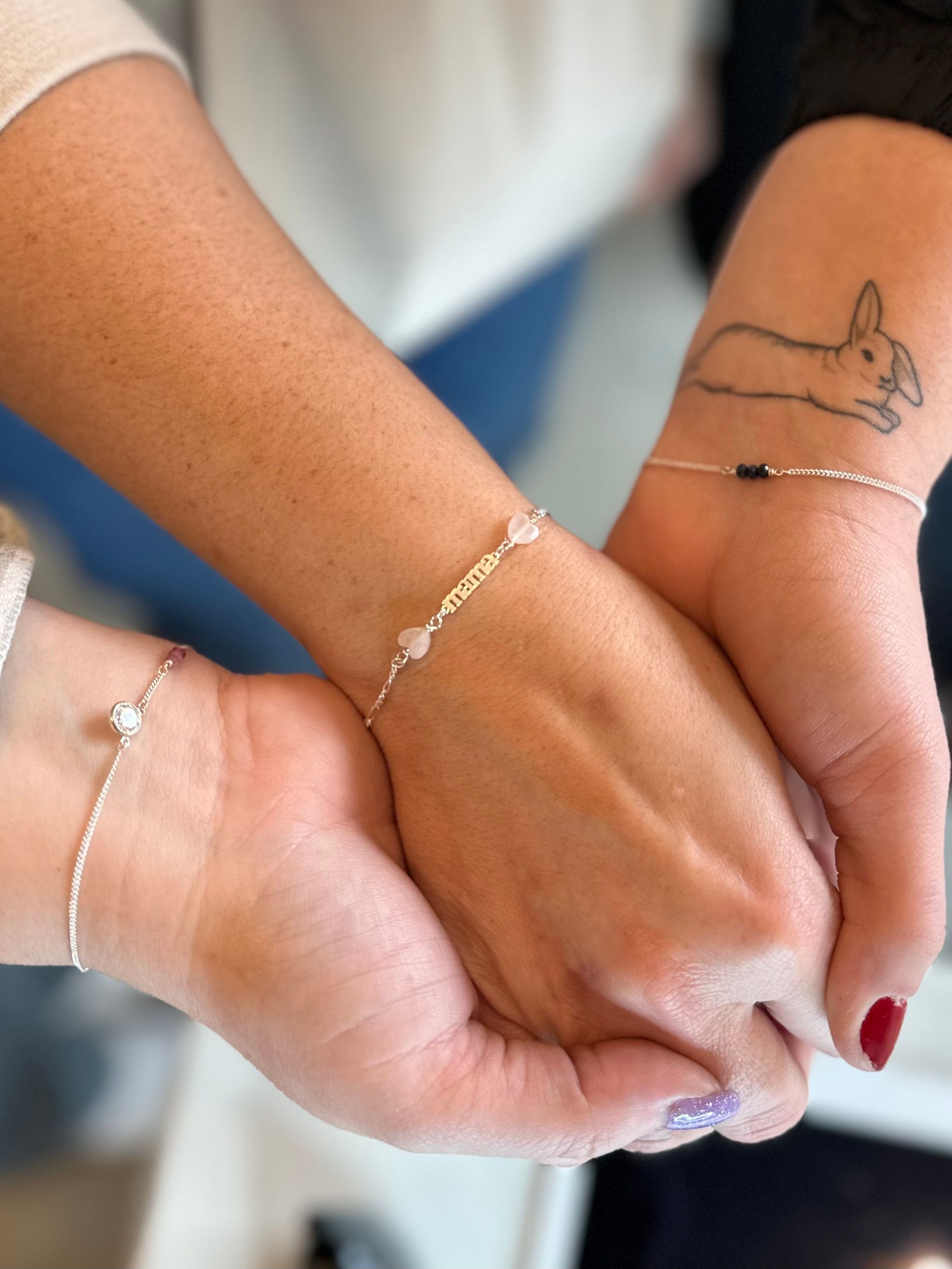 Close-up of three joined hands and wrists showcasing delicate silver bracelets and a nameplate with heart beads; one wrist has a rabbit tattoo and nails painted red and purple glitter, friendship-style jewelry shot.