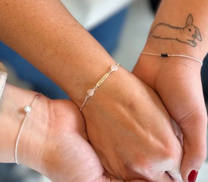 Close-up of three joined hands and wrists showcasing delicate silver bracelets and a nameplate with heart beads; one wrist has a rabbit tattoo and nails painted red and purple glitter, friendship-style jewelry shot.