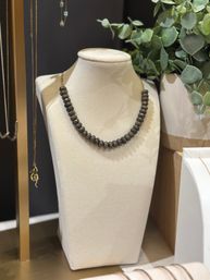 Dark metallic beaded necklace on a cream velvet display bust, styled beside eucalyptus greenery and hanging gold chains in a jewelry display.