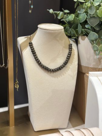 Dark metallic beaded necklace on a cream velvet display bust, styled beside eucalyptus greenery and hanging gold chains in a jewelry display.