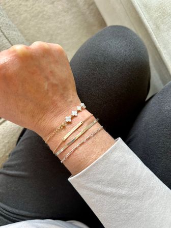 Close-up of a wrist adorned with stacked gold and silver bracelets — thin chains, hammered metal bars and small white clover beads — resting on a lap in dark leggings on a beige sofa.