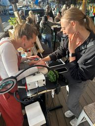 Pop-up outdoor manicure on a sunny downtown shopping street — nail technician filing a customer's nails at a sidewalk table with lamp, drill, tools and passersby in the background.