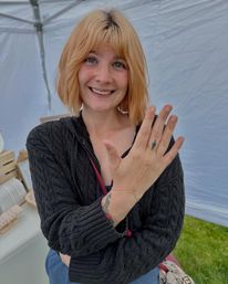 Smiling woman with short blonde hair and nose ring shows a delicate gold hand-chain bracelet and tattoos under a white market tent at an outdoor craft fair