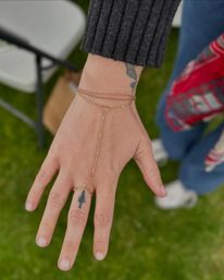 Close-up of a hand over green grass wearing a delicate gold hand-chain connecting a wrist bracelet to a ring, with a small pine-tree finger tattoo and a dark ribbed sleeve visible.