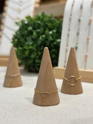 Close-up of a minimalist jewelry display with three tan cone ring holders showcasing delicate gold and silver chain rings, featuring triangle and circle designs against a blurred green plant backdrop.