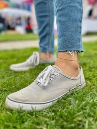 Close-up of gray canvas sneakers and a delicate gold anklet on a frayed-hem jean leg standing on green grass at an outdoor event