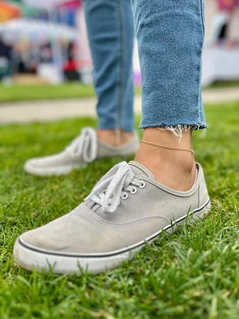 Close-up of gray canvas sneakers and a delicate gold anklet on a frayed-hem jean leg standing on green grass at an outdoor event