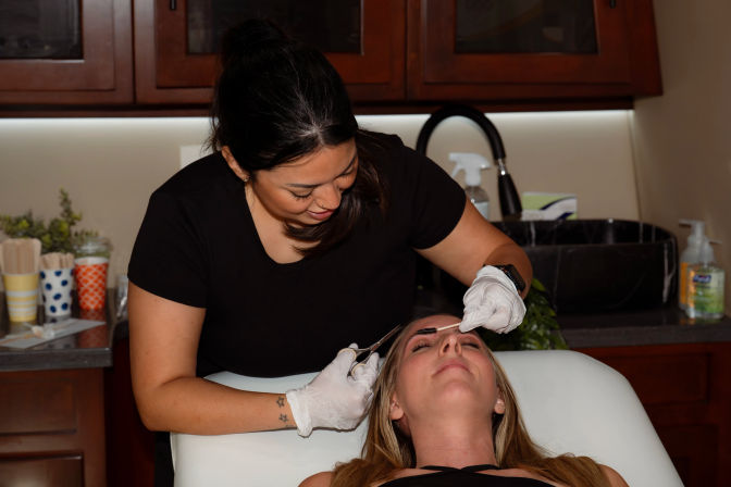 Expert brow shaping: esthetician in gloves grooms a client's eyebrows with a spoolie and scissors during a professional beauty salon treatment.
