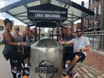 Smiling group of adults riding a pedal-powered party bike down a sunny downtown Birmingham, Alabama street lined with brick buildings