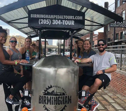 Smiling group of adults riding a pedal-powered party bike down a sunny downtown Birmingham, Alabama street lined with brick buildings