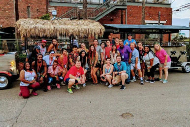 Large group of people smiling and posing beside a tiki-roofed pedal party vehicle on a city street with a red brick building backdrop