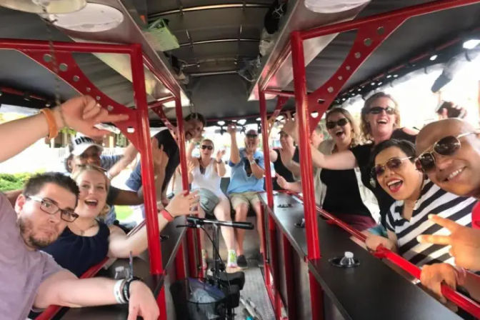 Smiling adults cheering aboard a red pedal-pub party bike on a sunny city street tour